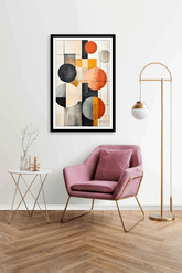 A modern geometric canvas painting featuring circles in black, white, gray, and orange, displayed in a room with a pink armchair.
