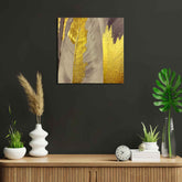 A canvas print featuring abstract gold and gray feathers displayed above a wooden frame on a dark wall, flanked by decorative plants and a clock on a wooden shelf.