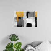 A canvas print featuring abstract geometric shapes in black, white, gray, and yellow, displayed above a grey sofa with a plant to the left.