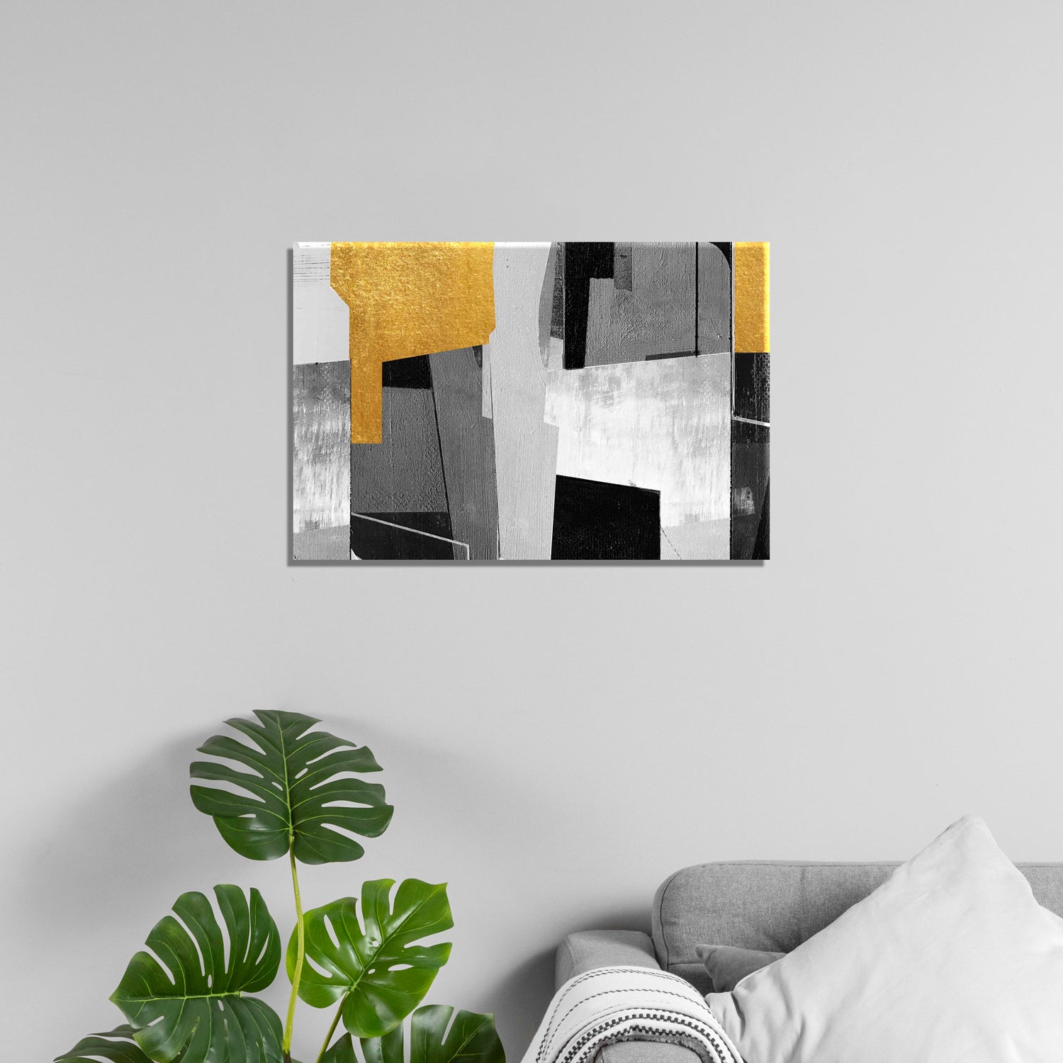 A canvas print featuring abstract geometric shapes in black, white, gray, and yellow, displayed above a grey sofa with a plant to the left.
